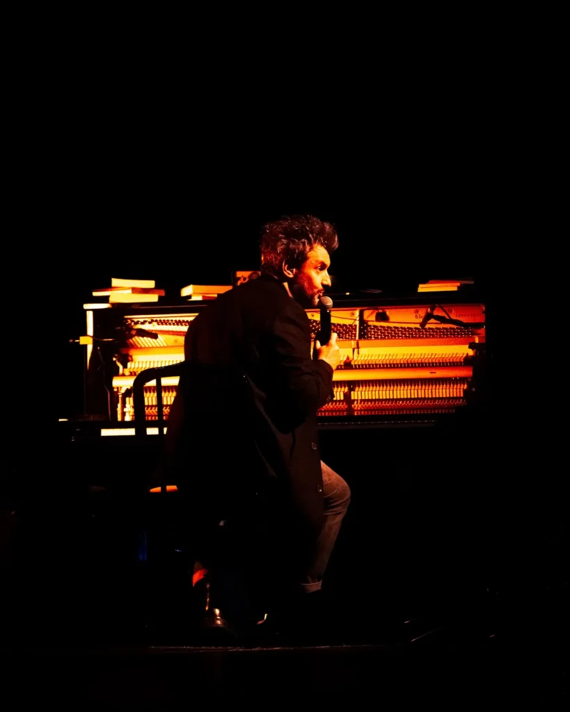 Rui Massena sentado de perfil em frente ao piano vertical aberto e iluminado a vermelho, segurando o microfone para falar com o público no concerto no Quartel das Artes em 2025, fotografia de concertos por xpectrum.