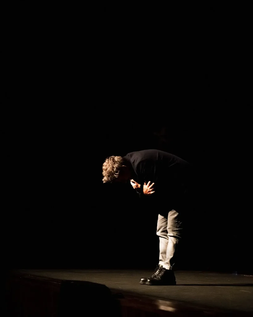 Rui Massena curvado em vénia ao público no final do concerto no Quartel das Artes em 2025, iluminado por um foco zenital sobre o palco escuro, fotografia de concertos por xpectrum.