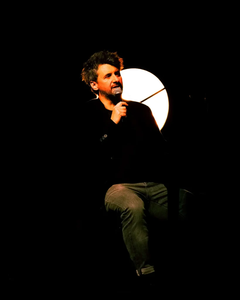 Rui Massena sentado de frente com microfone na mão a falar para o público, recortado por um foco circular branco quente atrás de si, durante o concerto no Quartel das Artes em 2025, fotografia de concertos por xpectrum.