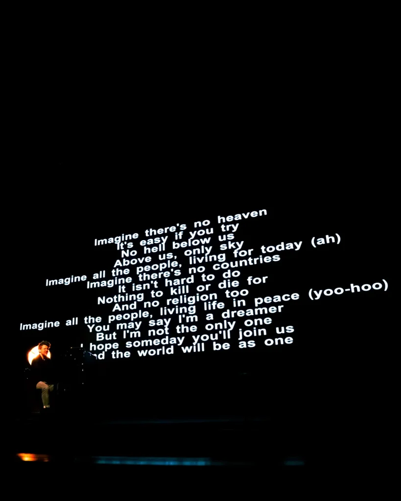 Letra de “Imagine” de John Lennon projetada em palco, com Rui Massena ao piano em pequena escala no canto inferior esquerdo, durante o concerto no Quartel das Artes em 2025, fotografia de concertos por xpectrum.