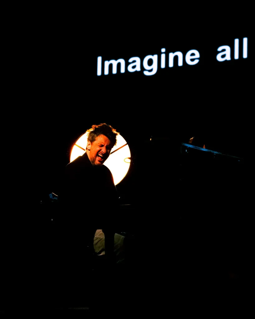 Rui Massena ao piano em expressão intensa e cantando, com as palavras “Imagine all” projetadas atrás de si, durante o concerto no Quartel das Artes em 2025, fotografia de concertos por xpectrum.