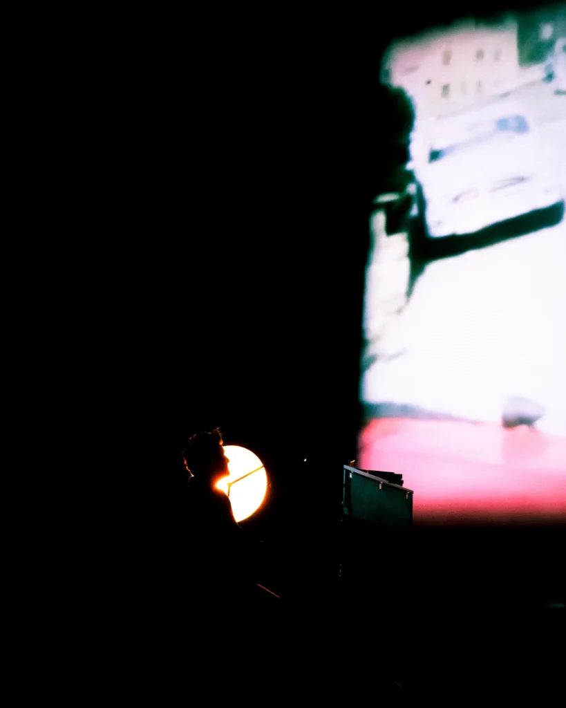 Silhueta lateral de Rui Massena ao piano recortada pelo candeeiro de palco, com vídeo projetado em tons rosa e branco no fundo do palco do Quartel das Artes em 2025, fotografia de concertos por xpectrum.